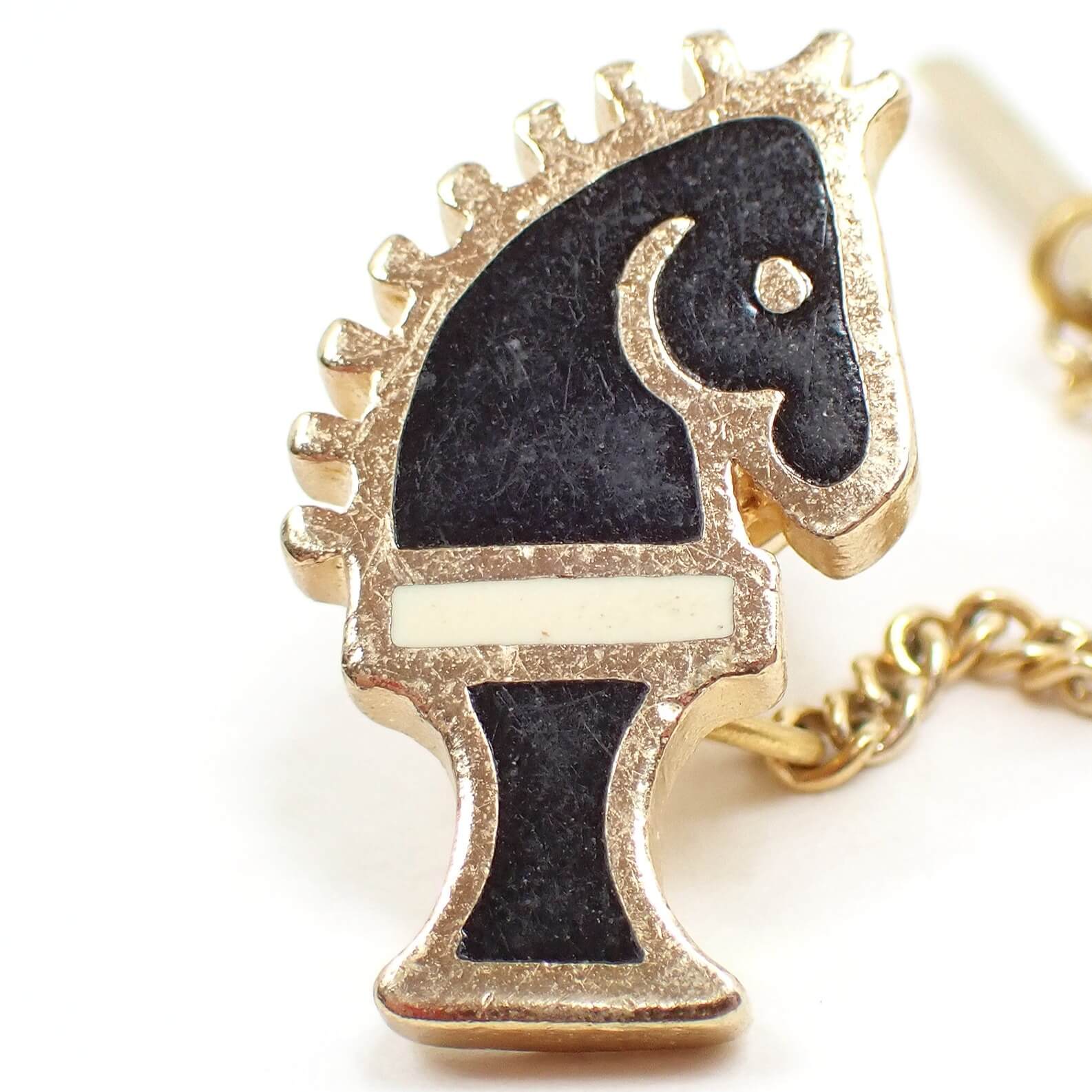 Enlarged front view of the retro vintage chess piece tie tack. The metal is gold tone in color. It is shaped like a knight horse chess piece. The main part of it is black enameled with an off white cream color stripe going across the middle.