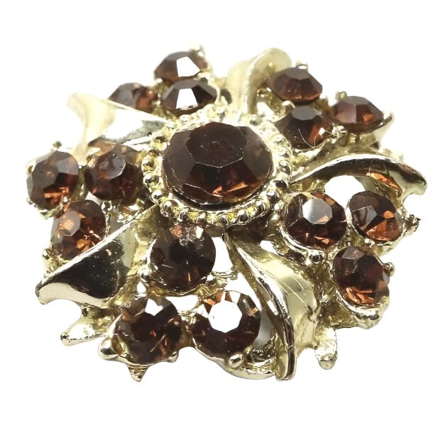 Front view of the Mid Century vintage rhinestone brooch pin. The metal is gold tone in color and has a starburst sort of shape. There is a brown rhinestone in the middle with smaller sized brown rhinestones around it in groups of three.