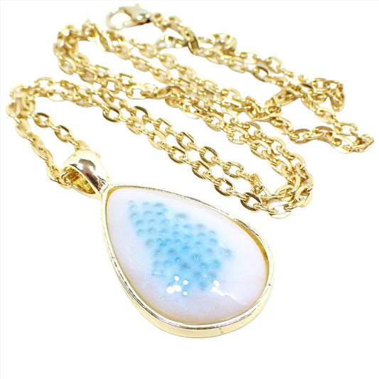 Angled view of the handmade resin teardrop pendant necklace. The front of the teardrop pendant has a domed resin cab that is white with a pearly sheen of blue. There are small blue bubble beads embedded in the middle area of the resin. The metal is gold tone in color but there are three dots in the lower right corner showing options for rose gold plated, gold plated, or silver plated.