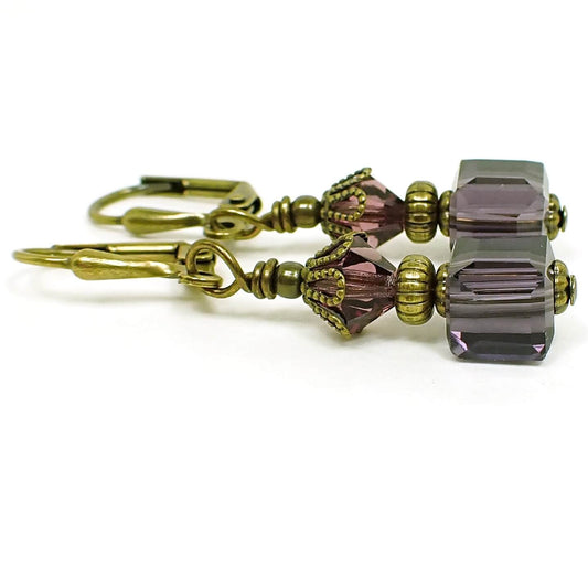 Side view of the small purple glass beaded earrings. The metal is antiqued brass in color. There are faceted glass crystal bicone beads at the top and small cube shaped glass beads at the bottom. The beads are purple in color.