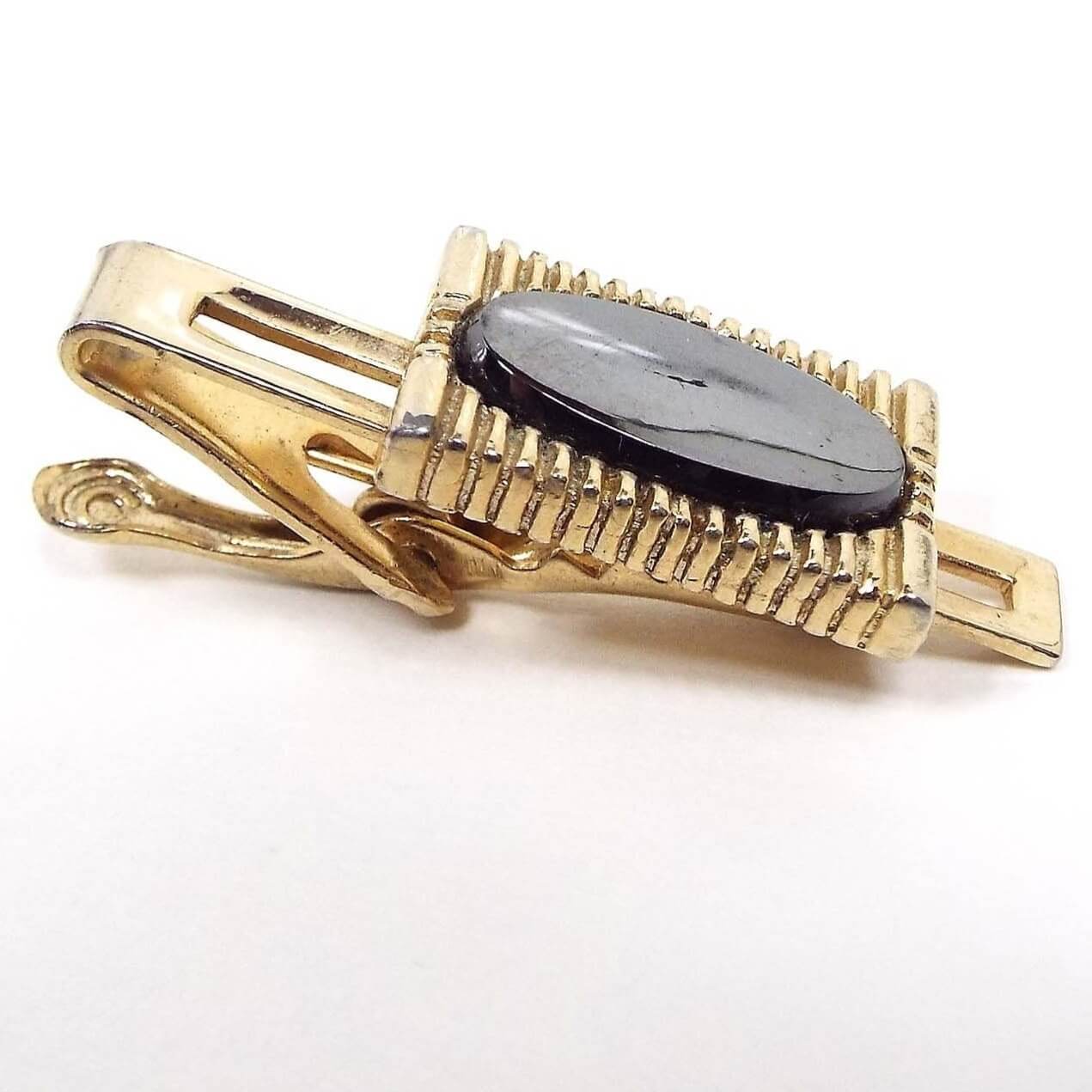 Angled front and side view of the Mid Century vintage hematite tie clip. The metal is gold tone in color. There is a striped rectangle in the middle with an oval hematite cab.