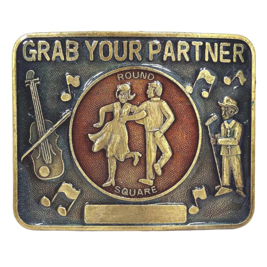 Front view of the retro vintage square dancing belt buckle. The metal is antiqued brass in color. It has a blue enameled area with Grab Your Partner, musical notes, and a square dancing caller. In the middle is a red enameled area that says Round Square with a couple dancing. There is a plain rectangle plate at the bottom.