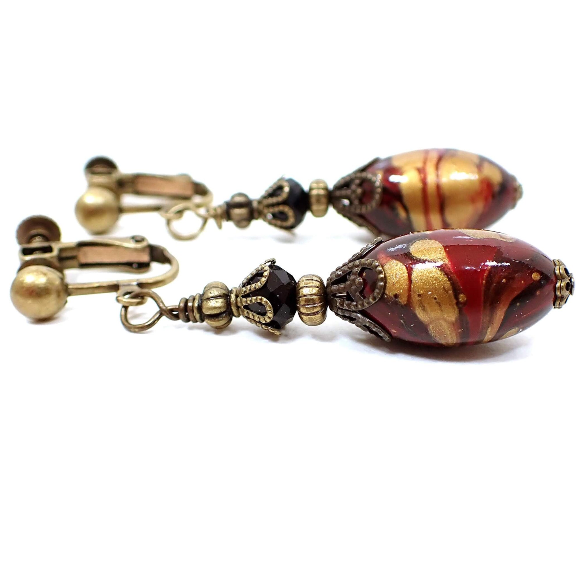 Side view of the handmade marbled lucite oval drop earrings. The metal is antiqued brass in color. There are black faceted glass beads at the top. The bottom beads are oval in shape and have marbled swirls of red, black, and antiqued gold color. Each lucite bead differs from the other for a unique appearance.