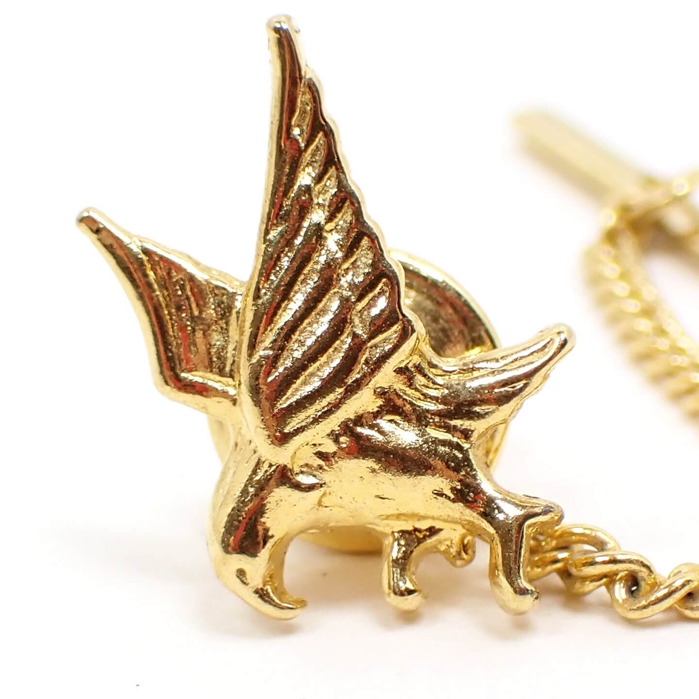 Enlarged front view of the retro vintage eagle tie tack. The metal is gold tone plated in color. The tie tack is shaped like a flying eagle with its sings out and talons spread to grab something or land.
