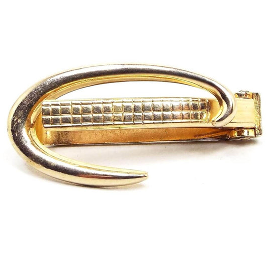 Front view of the retro vintage Modernist style tie clip by Swank. It is gold tone in color and has a curved almost C shaped design. There is a textured bar on the front with a waffle pattern that pushes in so the curve goes over the front of the tie.