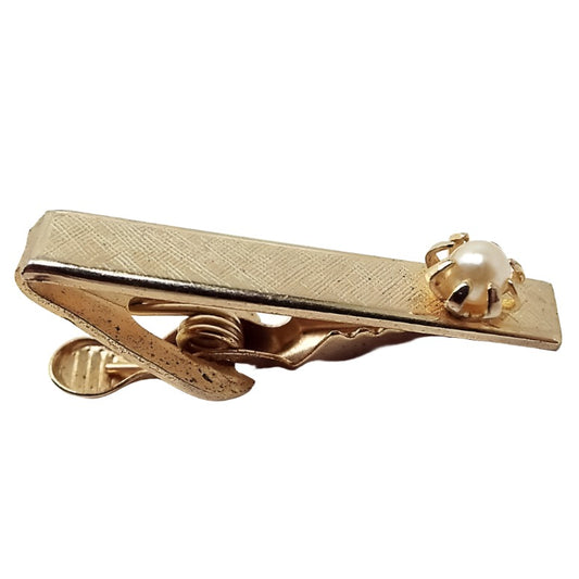 Front view of the Mid Century vintage faux pearl tie clip. The metal is gold tone in color. There is a prong set light yellow off white plastic imitation pearl at the end.