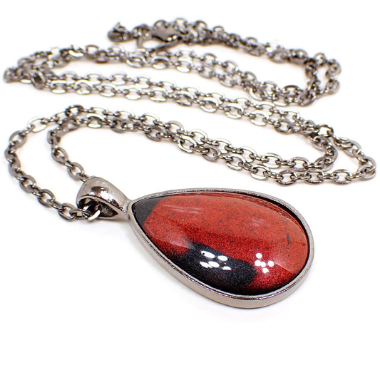 Angled view of the handmade resin teardrop pendant. The chain and setting and gray gunmetal plated. The  pendant is teardrop shaped with a domed resin cab. The resin is shimmery bright red in color and has a few splashes of black here and there.