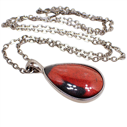 Angled view of the handmade resin teardrop pendant. The chain and setting and gray gunmetal plated. The  pendant is teardrop shaped with a domed resin cab. The resin is shimmery bright red in color and has a few splashes of black here and there.