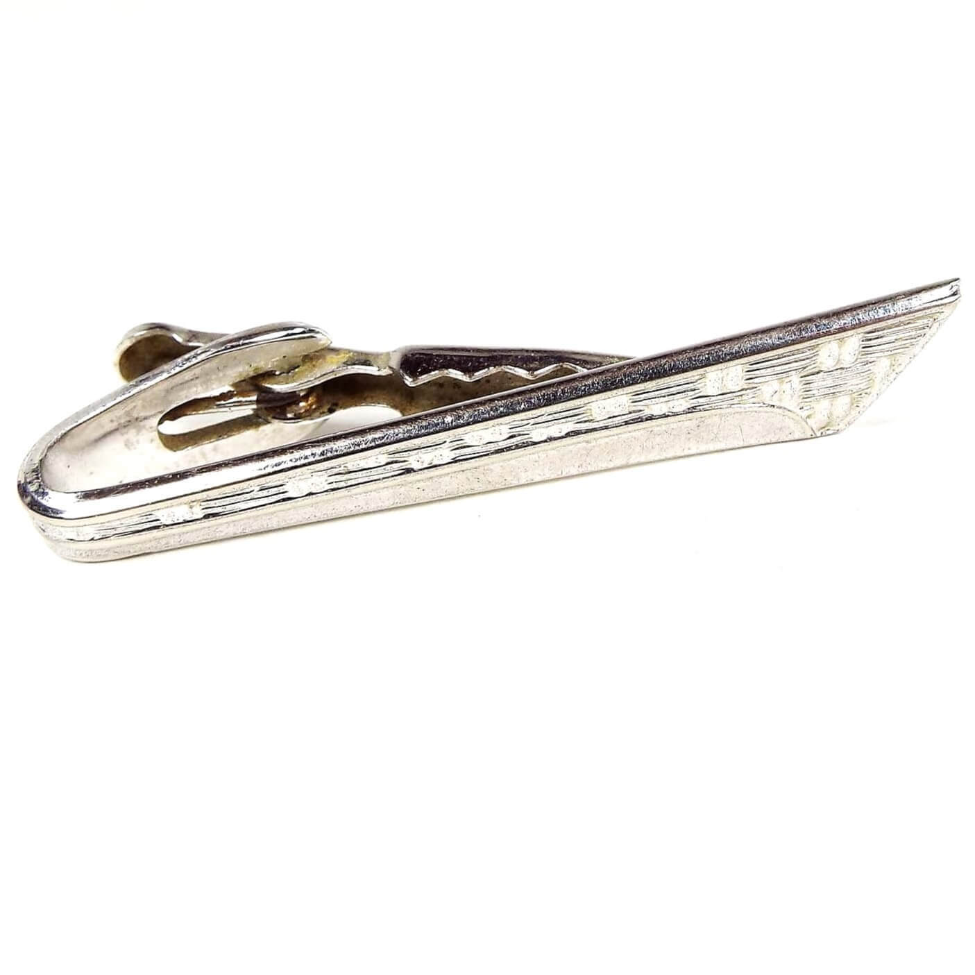 Front view of the Mid Century vintage Shields Modernist style tie clip. It is silver tone in color and has a textured pattern on the top part of the front with a thin curve design on the bottom. The end is angled and flares out at the top. 