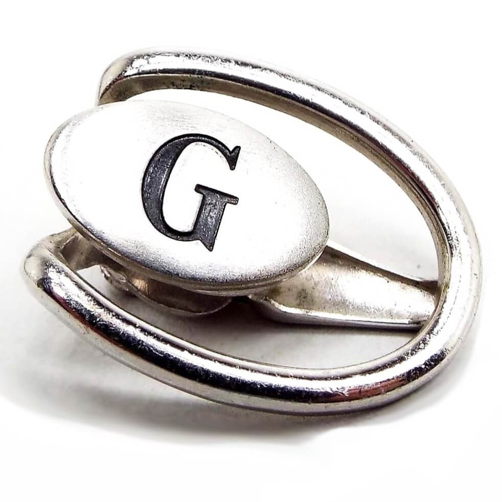 Enlarged front view of the Mid Century vintage Swank initial tie clip. It is silver tone in color and shaped like an open oval with a solid oval in the middle. The middle oval has the letter G engraved on it and is painted black that is worn to a dark gray in color from age.
