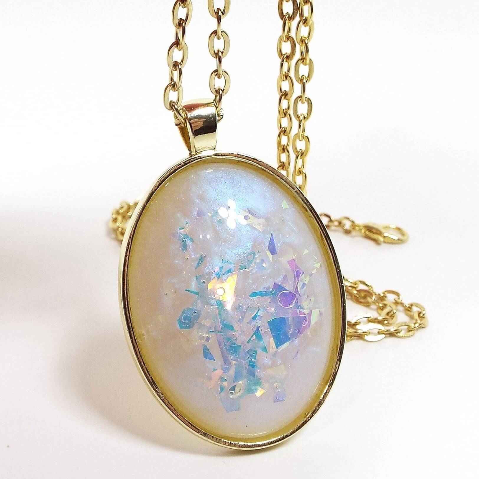 Front view of the handmade oval resin pendant necklace. The metal is gold tone in color. The oval pendant has a domed cab with color shift resin that has shades of pearly off white and blue. There are large chunks of AB glitter with blue, purple, and pink hues showing inside the resin for flashes of color.