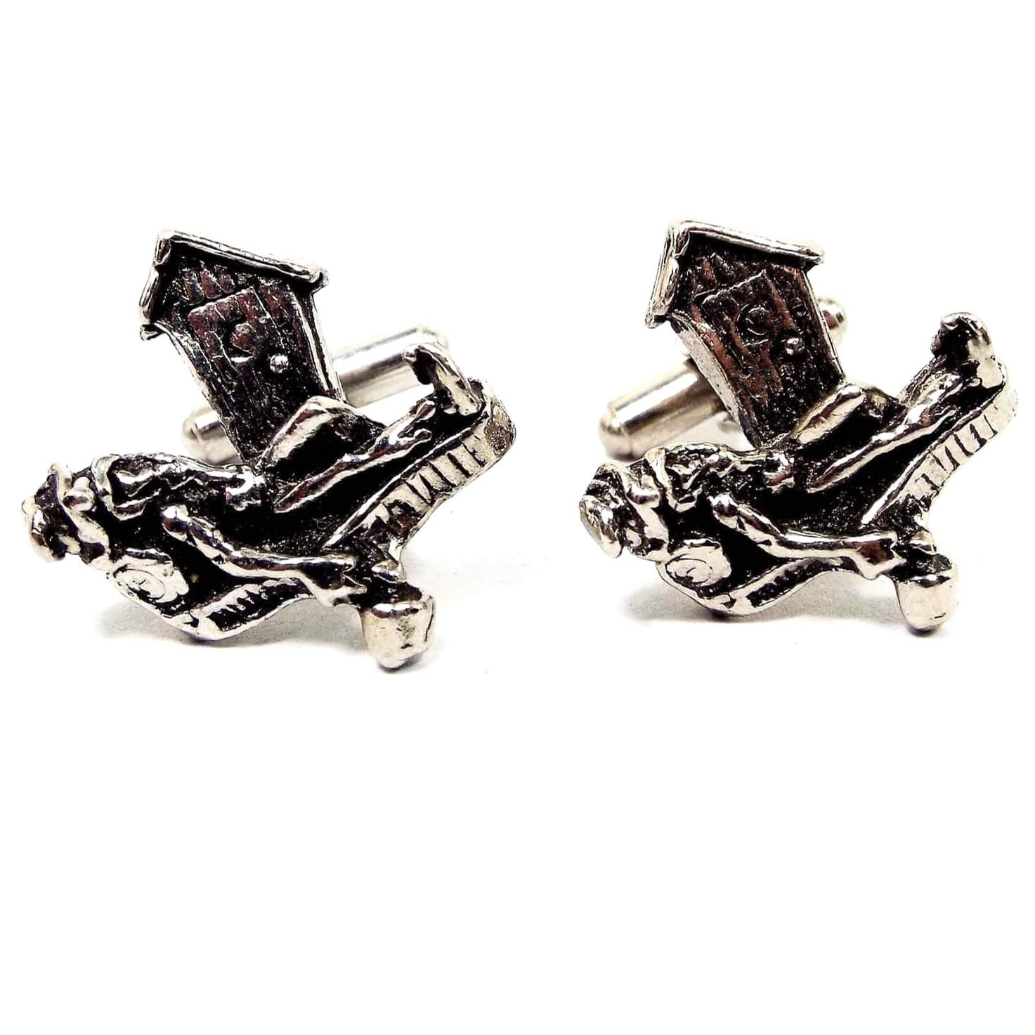 Front view of the Mid Century vintage Hillbilly cufflinks. They are antiqued silver tone in color. There is a mountain man type person with beard and mustache and hat laying on the front with a bedroll holding his jug of spirits. He is laying in front of an outhouse.