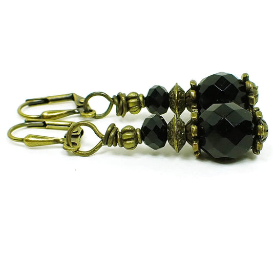 Side view of the Goth black glass drop handmade earrings. The metal is antiqued brass in color. There is a faceted rondelle shaped black bead at the top and a faceted oval black bead at the bottom. 