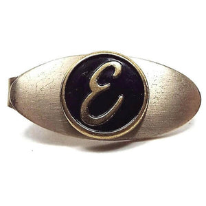 Front view of the retro vintage initial tie clip. It is a small oval that's gold tone in color and has a black enameled circle in the middle with the letter E in it.