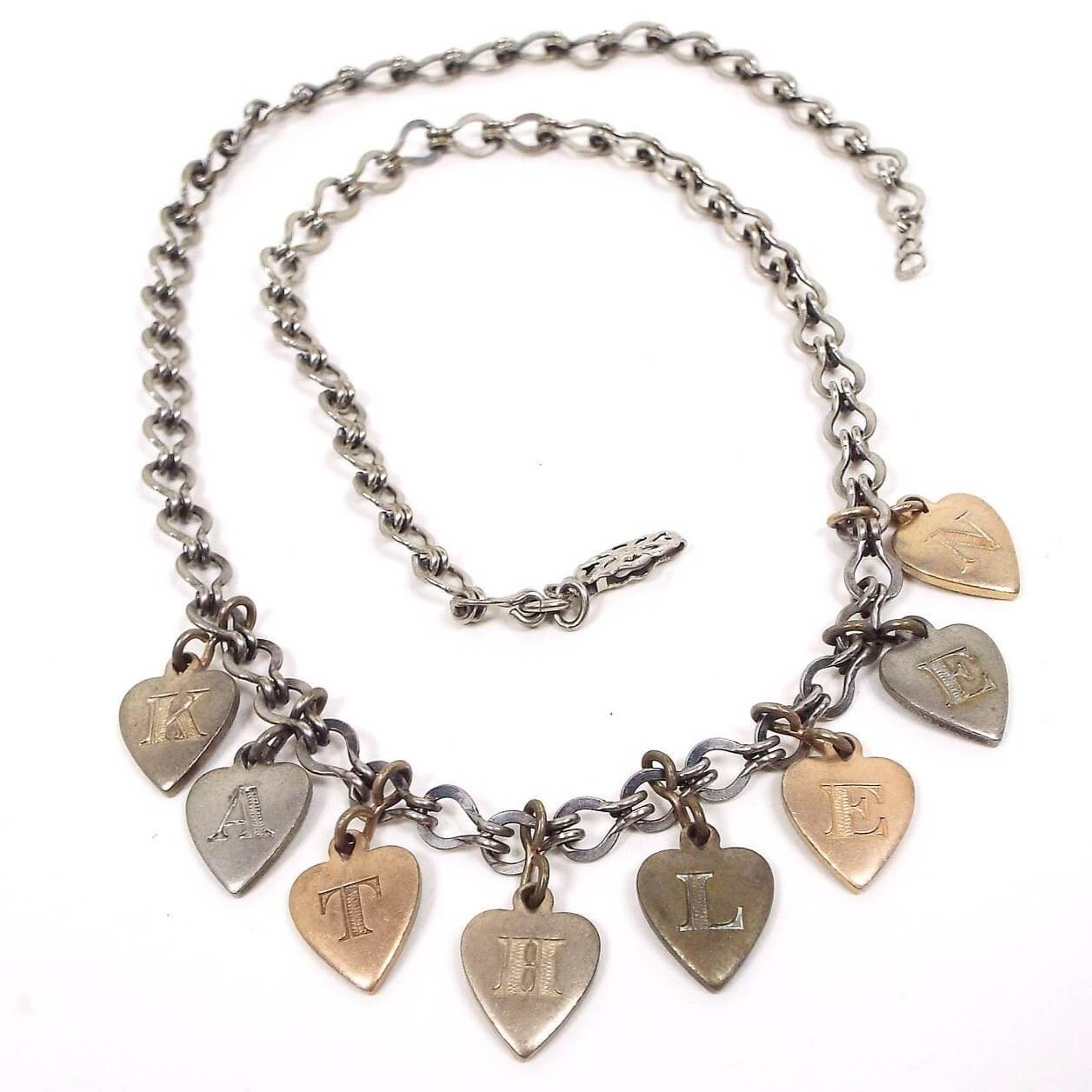 Front view of the Mid Century vintage heart charm name necklace. The chain has fancy style links and a filigree hinge clasp at the end. There are heart charms hanging from the bottom in different metal colors and each one has a letter engraved on it. All together they spell out the name Kathleen.