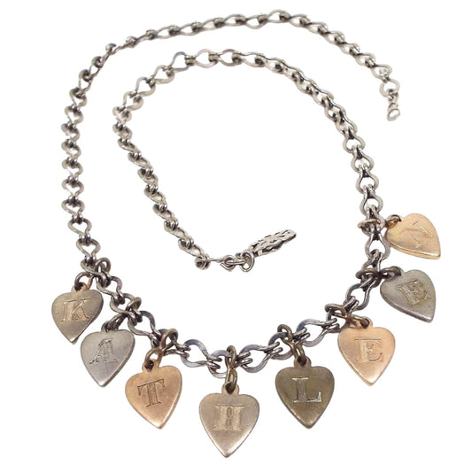 Front view of the Mid Century vintage heart charm name necklace. The chain has fancy style links and a filigree hinge clasp at the end. There are heart charms hanging from the bottom in different metal colors and each one has a letter engraved on it. All together they spell out the name Kathleen.