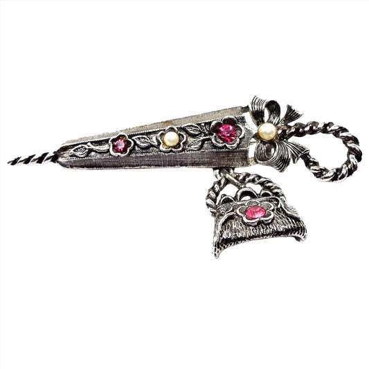 Front view of the retro vintage pewter brooch pin. It is shaped like an umbrella with a bow and has a dangling purse charm underneath it. There are flower shapes with fuchsia pink purple rhinestones in the middle and another flower shape with a faux pearl in the middle. There is also a faux pearl in the middle of the bow on the umbrella handle.