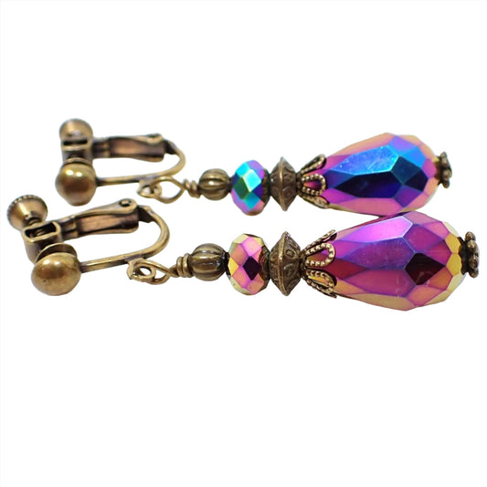 Side view of the handmade rainbow metallic multi color earrings. They have saucer shaped faceted glass beads at the top and faceted glass teardrop beads at the bottom. They colors are different as you rotate around the beads in pink, blue, purple, and yellow. The metal is antiqued brass in color.
