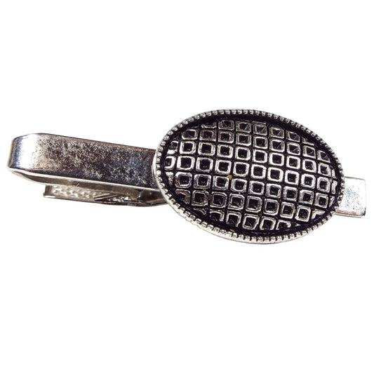 Front view of the retro vintage Judy Lee tie clip. It is silver tone in color and has an oval at the end. The inside of the oval has a pattern of tiny squares with black around the edge and middle of the squares.