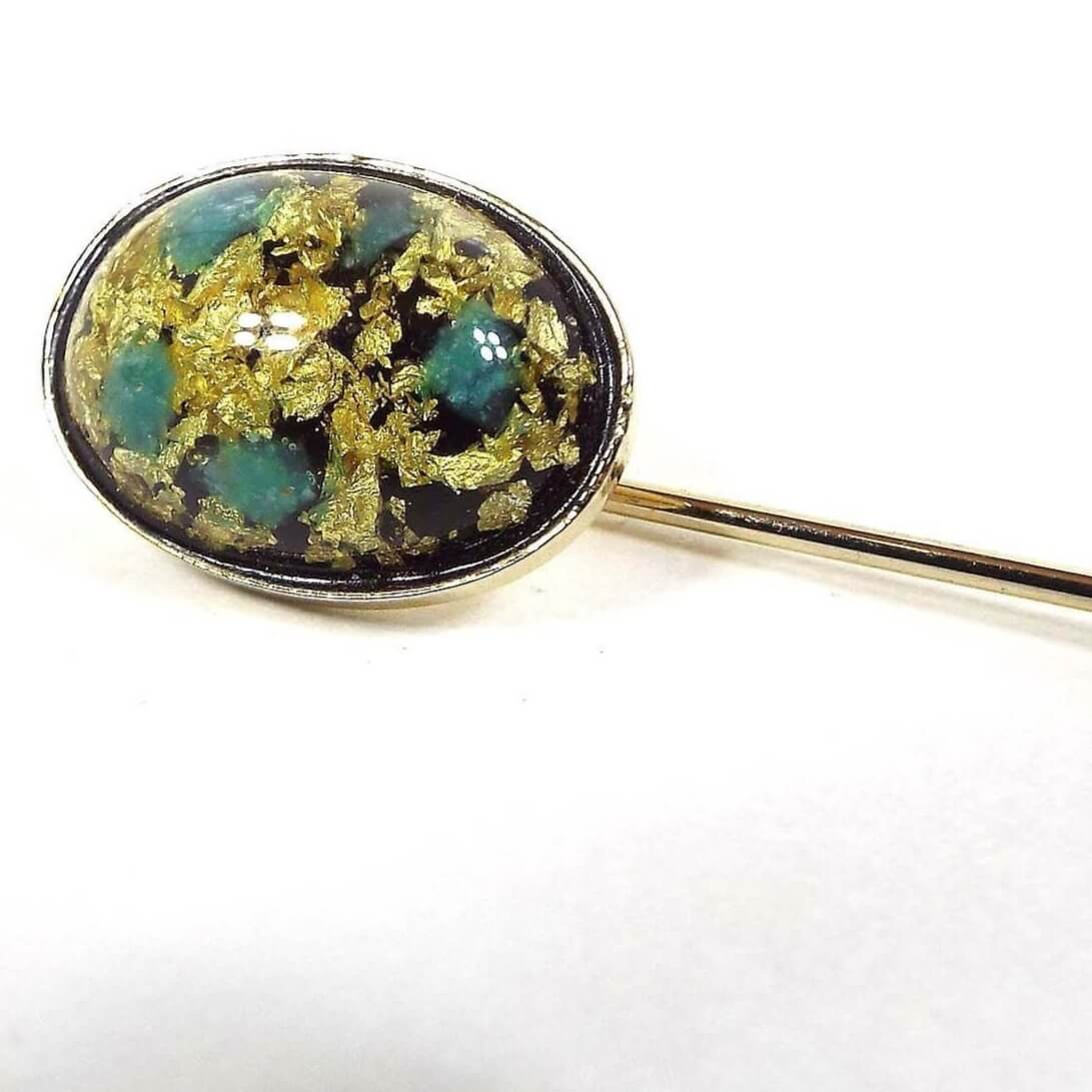 Front view of the Mid Century vintage confetti lucite stick pin. the metal is gold tone in color. There is an oval lucite cab at the top with gold color flakes and spots of mint green and blue.