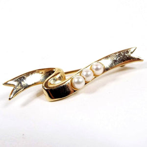 Front view of the 1984 Avon President's Club brooch pin. It is gold tone in color and shaped like a curved ribbon. There are three imitation glass pearls on the front.