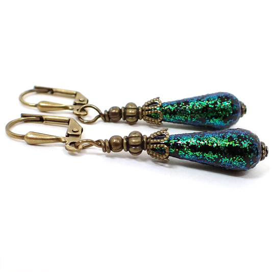 Angled side view of the handmade small teardrop earrings. They have antiqued brass color metal with small black vintage acrylic teardrop beads at the bottom that have bright green glitter embedded in them.