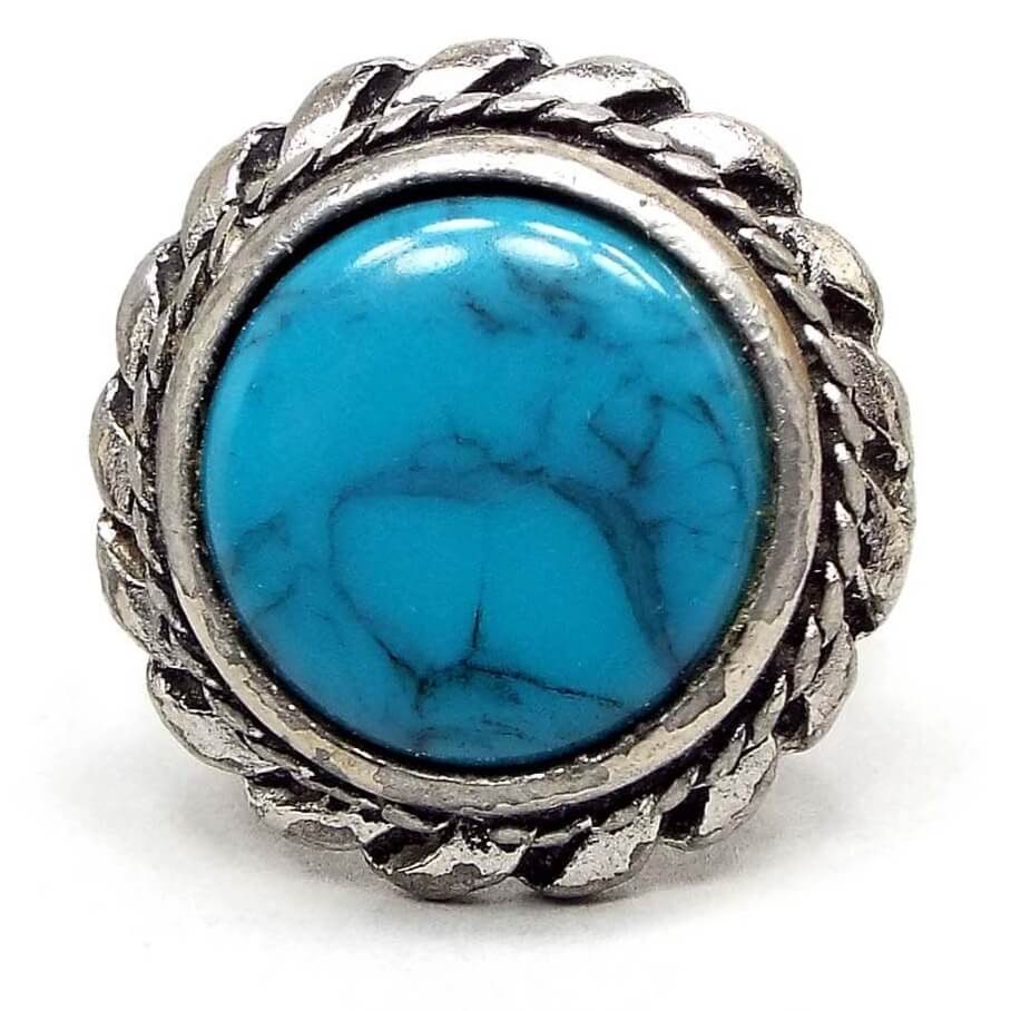 Front view of the retro vintage Southwestern style faux turquoise ring. The front setting is silver tone pewter and has a braided rope design on the outside edge of the bezel. The outer edge has a sort of short pinwheel style design. The middle cab is blue with gray and black lines marbled in. Chips from the silver plating can be seen on the setting and edges.
