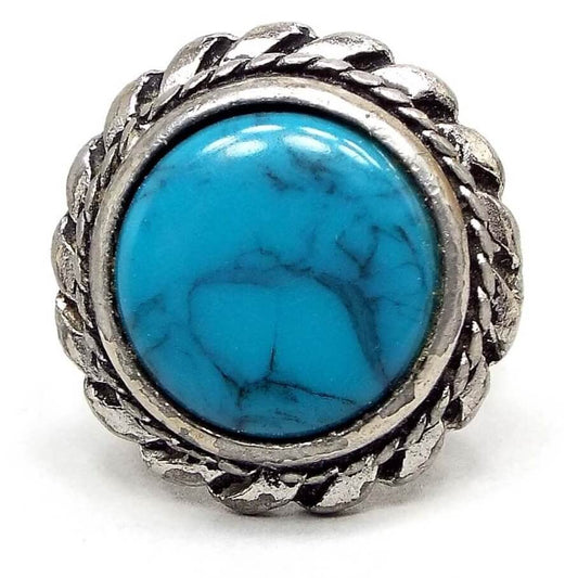 Front view of the retro vintage Southwestern style faux turquoise ring. The front setting is silver tone pewter and has a braided rope design on the outside edge of the bezel. The outer edge has a sort of short pinwheel style design. The middle cab is blue with gray and black lines marbled in. Chips from the silver plating can be seen on the setting and edges.