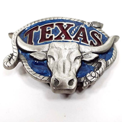Front view of the retro vintage Texas belt buckle by Great American Buckle Company. It is light pewter gray in color with blue enamel back ground,. There is a steer on the front and the edge has a rope design. It has Texas at the top in red enamel. 