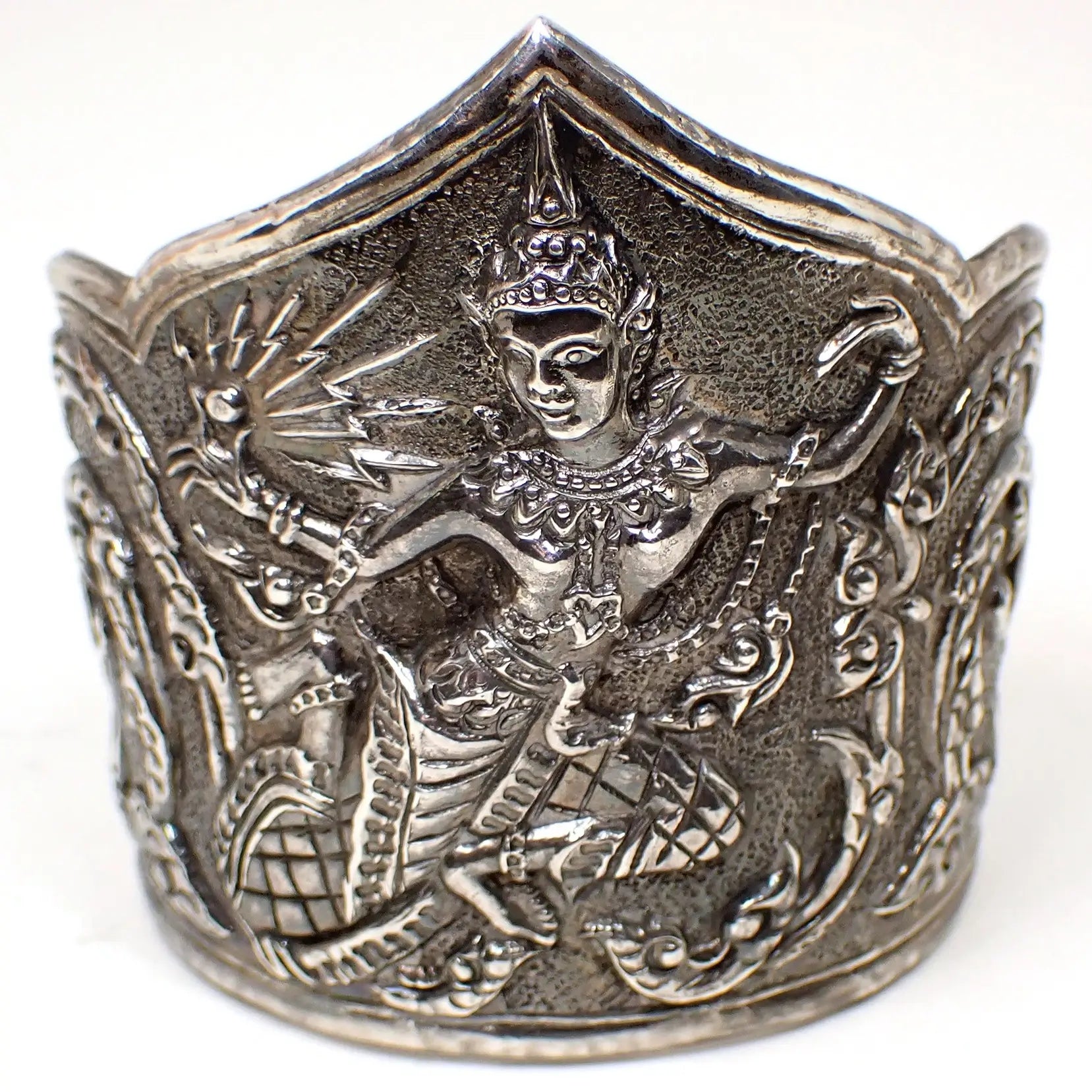 Front view of the Mid Century vintage Thai cuff bracelet. The sterling silver is a darkened gray in color. The bottom edge is rounded and the top edge has a point in the middle and rounded edges on the sides. There is a raised repousse design of a Siam dancer on the front with vines and leaves on either side. 