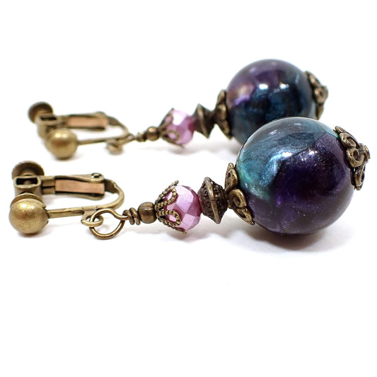 Side view of the handmade drop earrings with vintage lucite beads. The metal is antiqued brass in color. The top beads are faceted glass in a very light purple color. The bottom lucite beads are around and marbled with pearly shades of green, purple, and blue, with a few hints of other colors. The vintage beads vary from one another for an interesting look.