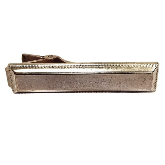 Front view of the Anson Mid Century vintage tie clip. The metal is a darkened gold tone in color from age. It has a rectangle design with a textured beveled edge.