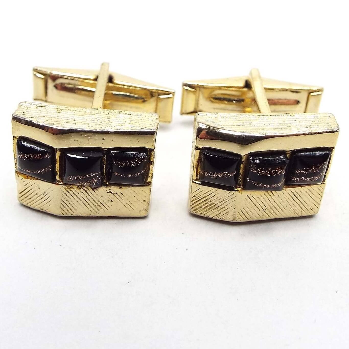 Front view of the retro vintage cufflinks with fancy glass cabs. The cufflinks are gold tone in color and are square with an angle on the front. There are three square glass cabs on each that are black with stripes of copper color glitter. No two cabs look the same. 