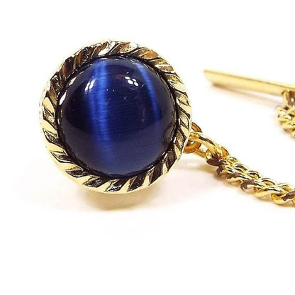 Enlarged front view of the retro vintage faux cat's eye tie tack. The metal is gold tone in color. The outer edge has a faceted design. There is a domed round dark blue glass cab in the middle with flashes of lighter blue as you move around in the light for a faux cat's eye appearance.