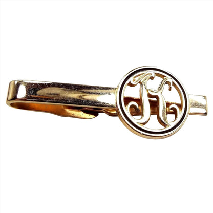 Front view of the retro vintage initial tie clip. It is gold tone in color. The end has an open cut out circle with the letter K inside and has black trim around the edge.