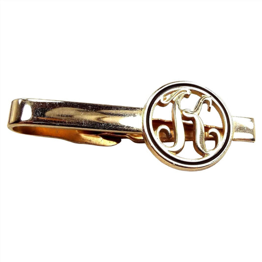 Front view of the retro vintage initial tie clip. It is gold tone in color. The end has an open cut out circle with the letter K inside and has black trim around the edge.