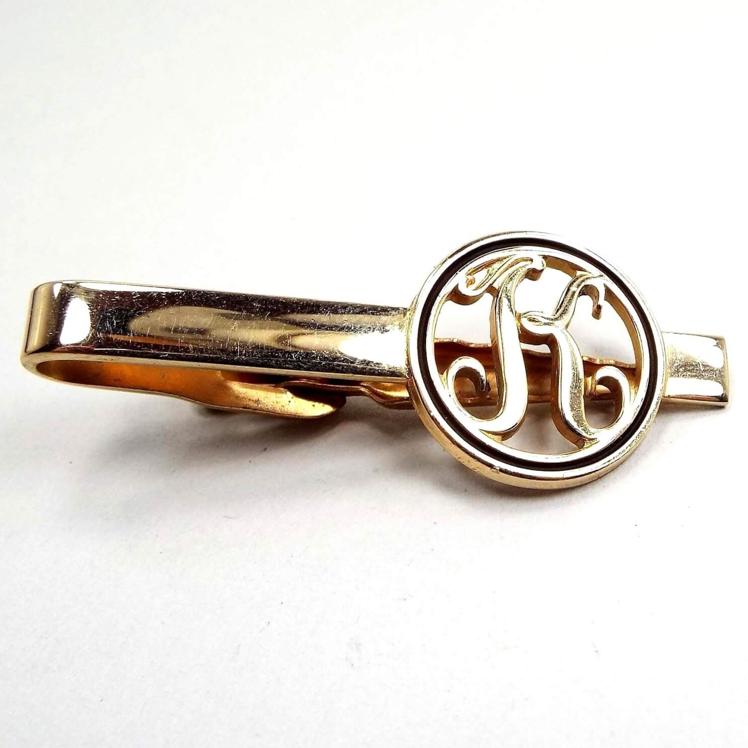 Front view of the retro vintage initial tie clip. It is gold tone in color. The end has an open cut out circle with the letter K inside and has black trim around the edge.