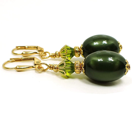 Side view of the handmade olive green oval drop earrings. The metal is gold plated in color. There is a faceted glass crystal olive green bead at the top and an oval moonglow lucite olive green bead at the bottom. The lucite bead has a glowy like appearance as you move around in the light.