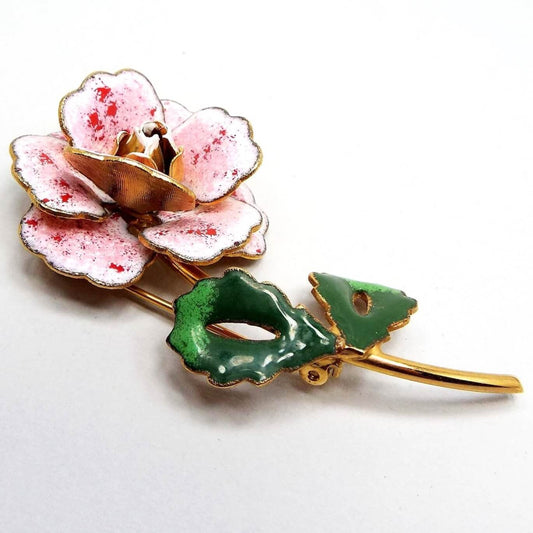 Front view of the retro vintage enameled floral brooch pin. The metal is gold tone in color. The brooch is shaped like a a flower with round scalloped petals that are enameled white and pink with red splotches on them. The leaves are open teardrop shaped and enameled in shades of green. The flower has a longer curved metal stem.