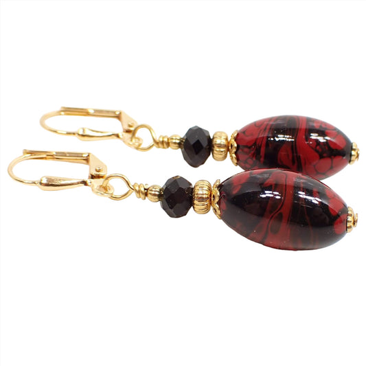 Side view of the handmade marbled oval lucite earrings. The metal is gold tone in color. There is a faceted black glass bead at the top and oval lucite beads at the bottom. The lucite beads are bright red swirled and marbled with black color. 