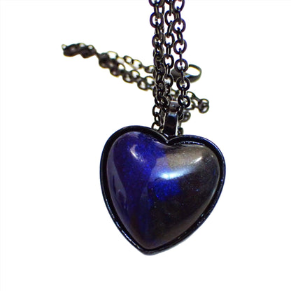 Enlarged front view of the Goth black heart handmade pendant necklace. The chain and pendant setting are black coated. The pendant is heart shaped with a domed handmade resin cab. The resin is pearly dark blue and black in color. 