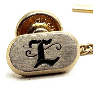 Enlarged front view of the Mid Century vintage Hickok initial tie tack. The front of the oval tie tack is silver tone in color with a black fancy script letter L on the front. The rest of the tie tack is gold tone in color.