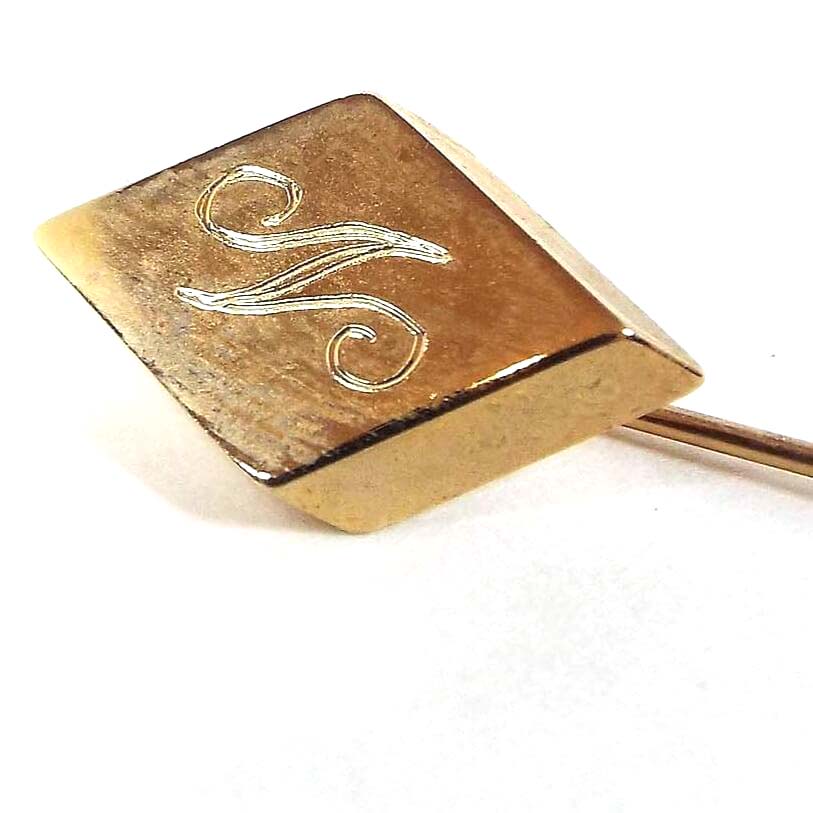 Enlarged view of the top of the Mid Century vintage initial stick pin. The metal is gold tone in color. There is a diamond shape at the top with the letter S engraved on it in fancy script.