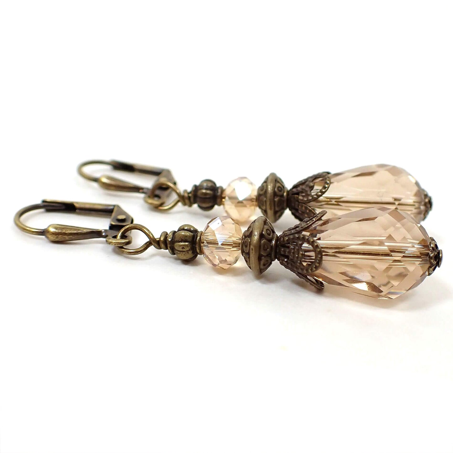 Side view of the vintage style light peach teardrop earrings. The metal is antiqued brass in color. There is a faceted glass crystal bead at the top and a faceted glass crystal teardrop bead at the bottom. 