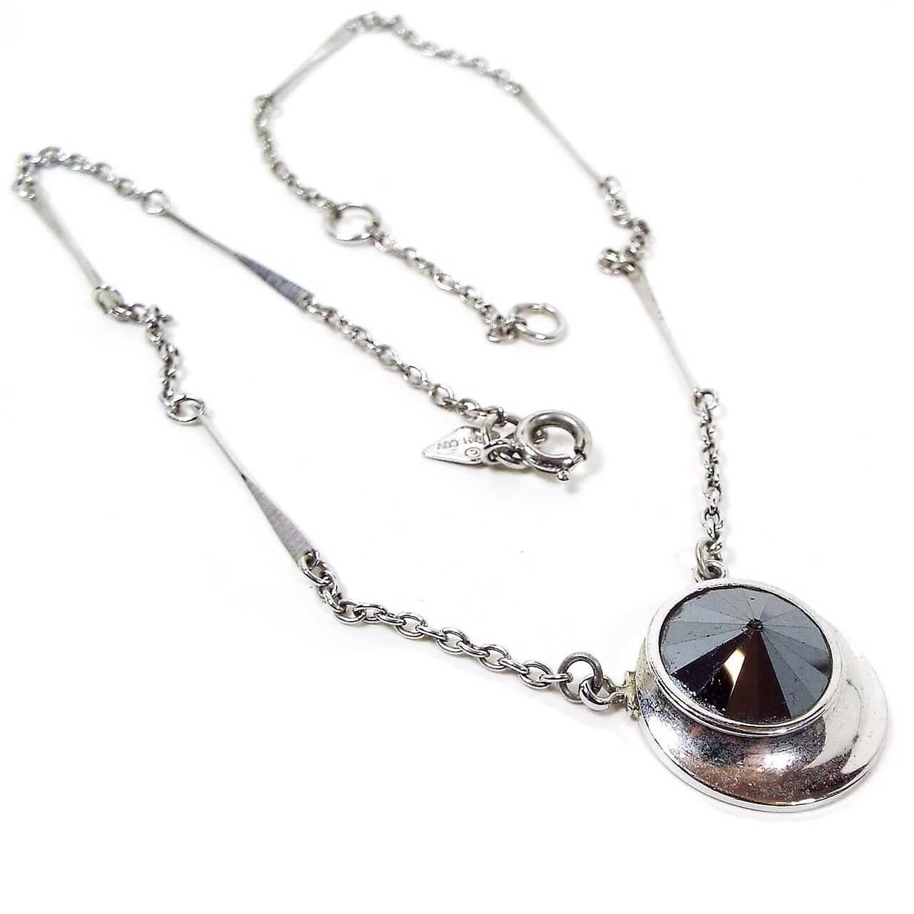Front view of the retro vintage Sarah Coventry pendant necklace. The metal is silver tone in color. The pendant is oval shaped with a round rivoli rhinestone at the top. The rhinestone is metallic gray in color for a faux hematite appearance. There is a spring ring clasp at the end with two rings on the chain to clasp it to for size. There is a Sarah Cov hang tag by the clasp.