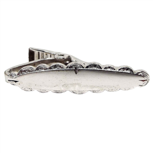 Front view of the retro vintage small tie clip. It is silver tone in color and oval shaped like a long surfboard. It has a textured scalloped shape all the way around the edge. 