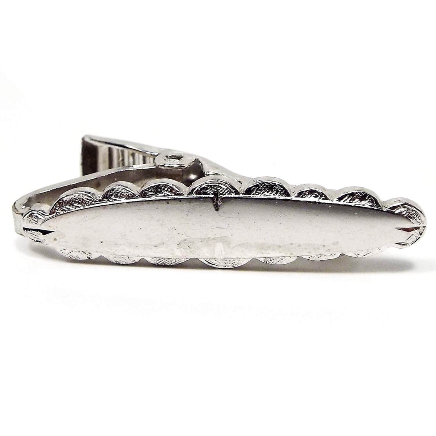 Front view of the retro vintage small tie clip. It is silver tone in color and oval shaped like a long surfboard. It has a textured scalloped shape all the way around the edge. 
