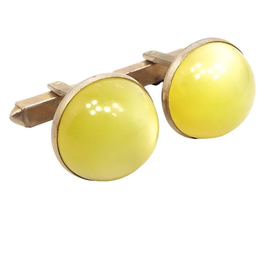 Angled front view of the 1950's Mid Century vintage moonglow lucite cufflinks. The metal is gold tone in color. The fronts are domed round shape with yellow moonglow cabs on the front.