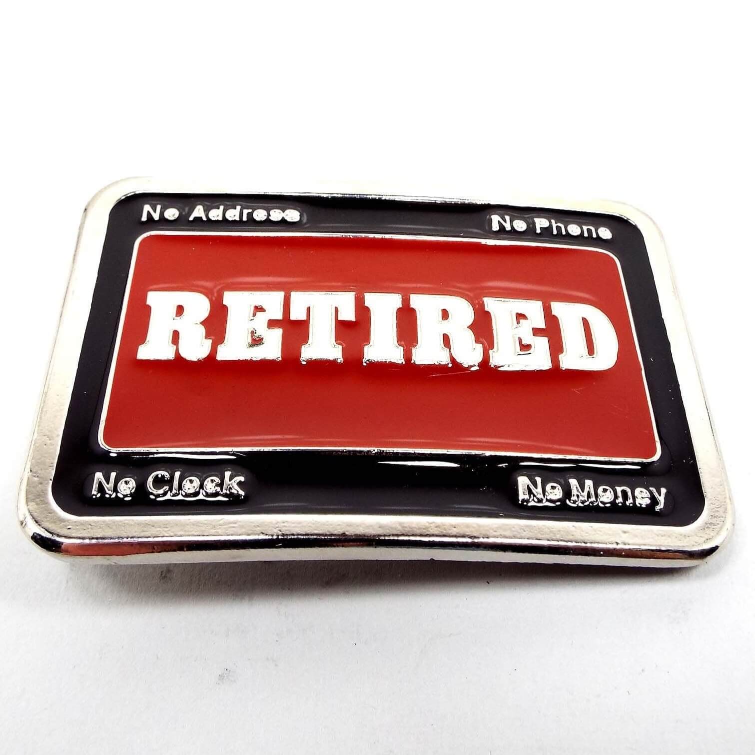 Front view of the retro vintage Retired belt buckle. The metal is silver tone in color. The middle says Retired with red enamel around it. The edge is black enameled and has no address, no phone, no clock, no money on the corners.