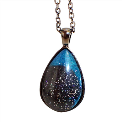 Enlarged front view of the handmade teardrop pendant necklace. The metal is gunmetal gray in color. The pendant has a handmade dome resin front with pearly dark gray and pearly teal blue areas of resin. The blue is primarily at the top of the pendant and the dark gray at the bottom. There is holographic silver glitter scattered around the resin for tiny flecks of sparkle and flashes of color.